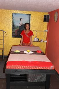 Darunee Goldammer Inhaberin von Thai Life Massage Velbert am Ende einer Massage-Liege, mit einem kunstvollen Gemälde im Hintergrund und Lotusblumenschmuck auf der Liege, der eine beruhigende Atmosphäre schafft.