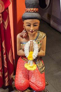 Nahaufnahme einer traditionellen thailändischen Holzfigur in thailändischer Tracht, frontal aufgenommen, die das thailändische ‚Wai‘ machend, eine respektvolle Geste des Begrüßens oder Betens symbolisiert, mit einem Blumenkranz um die Hände.