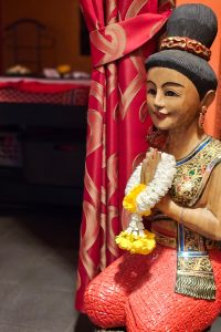 Nahaufnahme einer traditionellen thailändischen Holzfigur in thailändischer Tracht, kniend und das thailändische ‚Wai‘ machend, die eine respektvolle Geste des Begrüßens oder Betens symbolisiert, mit einem Blumenkranz um den Händen.