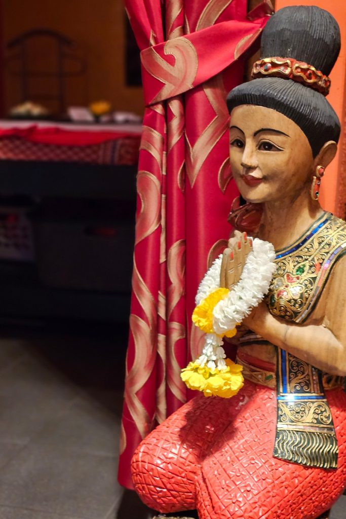 Nahaufnahme einer traditionellen thailändischen Holzfigur in thailändischer Tracht, kniend und das thailändische ‚Wai‘ machend, die eine respektvolle Geste des Begrüßens oder Betens symbolisiert, mit einem Blumenkranz um den Händen.