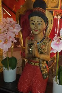 Nahaufnahme einer traditionellen thailändischen Holzfigur in thailändischer Tracht, kniend und das thailändische ‚Wai‘ machend, eine respektvolle Geste des Begrüßens oder Betens symbolisierend, im Schaufenster, umgeben von Orchideen.
